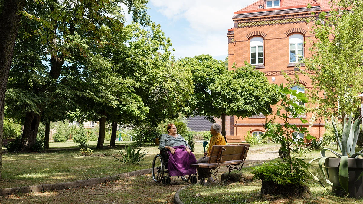 Dwie podopieczne rozmawiają w zielonym parku na terenie ośrodka Niebieski Parasol w Chojnowie – jedna siedzi na wózku inwalidzkim, a druga na ławce, w tle widoczny jest zabytkowy budynek z czerwonej cegły.