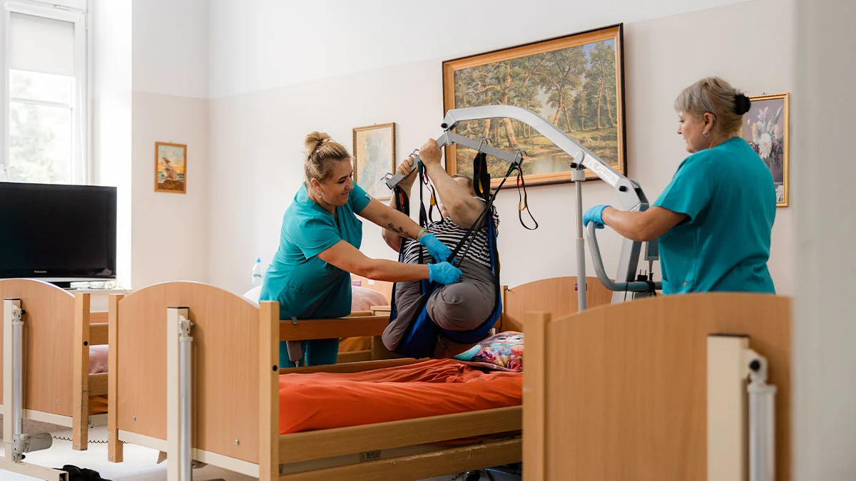 Dwie opiekunki w turkusowych strojach z ośrodka Niebieski Parasol pomagają pacjentowi przy użyciu podnośnika w sali z łóżkami rehabilitacyjnymi.