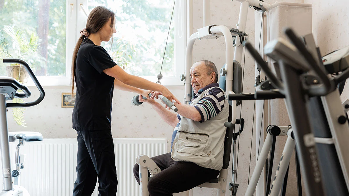 Fizjoterapeutka z ośrodka Niebieski Parasol asekuruje starszego mężczyznę ćwiczącego na wielofunkcyjnym atlasie treningowym na sali rehabilitacyjnej. Fizjoterapeutka z ośrodka Niebieski Parasol asekuruje starszego mężczyznę ćwiczącego na wielofunkcyjnym atlasie treningowym na sali rehabilitacyjnej.